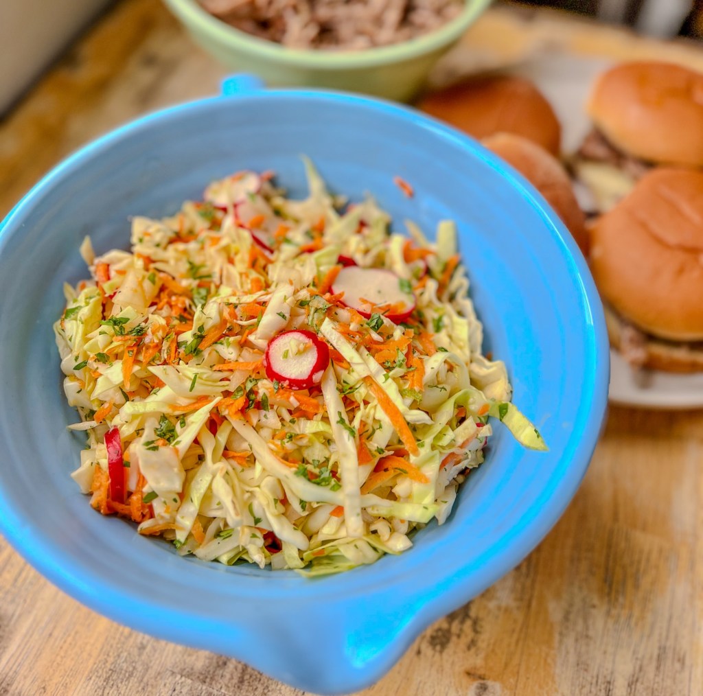 Cabbage slaw in a blue bowl with some pulled pork sandwiches behind it