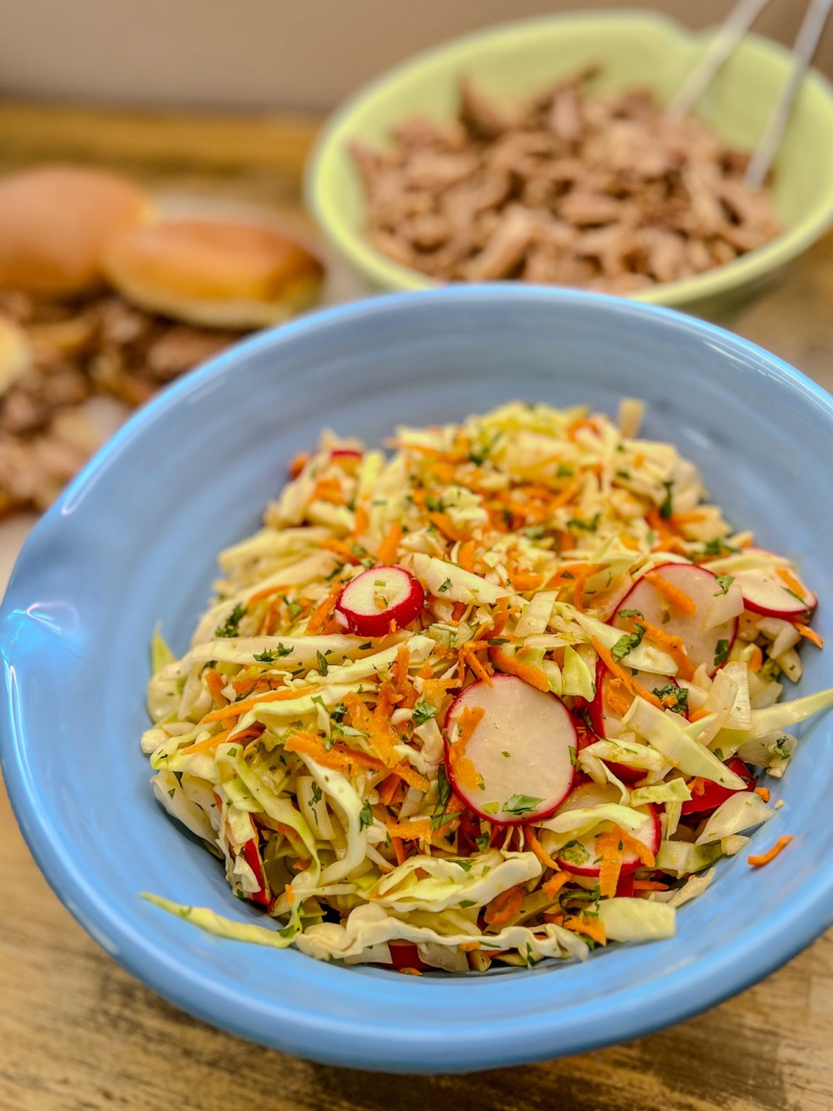 A big blue bowl filled with cabbage slaw with some pulled pork and sandwiches behind on a wooden table