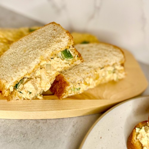 A cajun chicken salad sandwich with some chips next to it on a wood board with a plate next to it