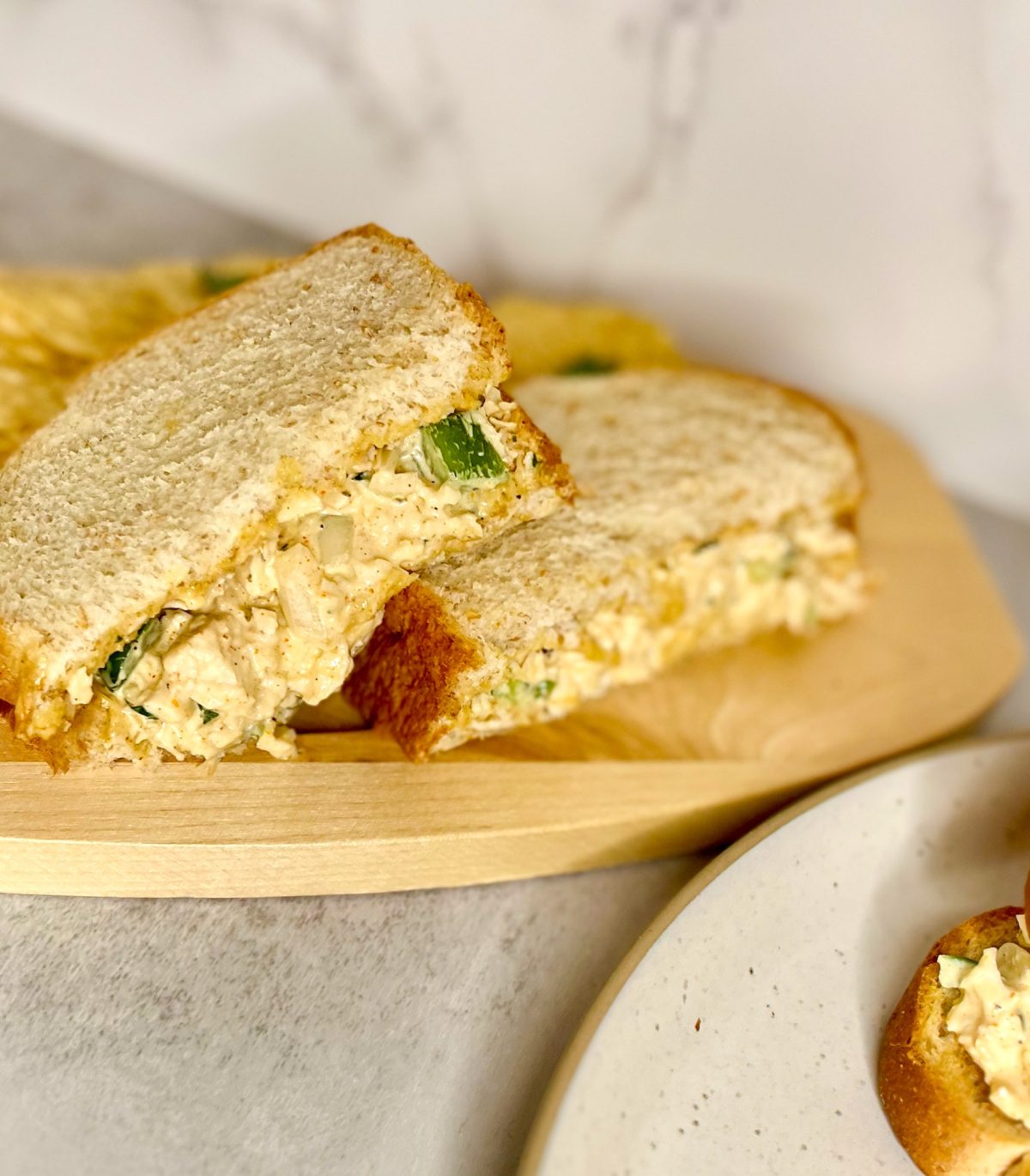 A cajun chicken salad sandwich with some chips next to it on a wood board with a plate next to it