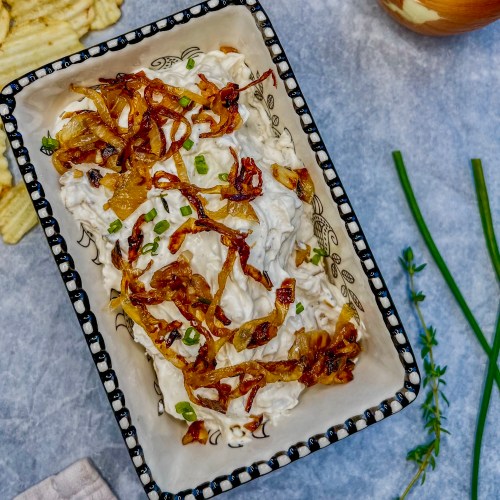Caramelized french onion dip in a dish with some chips, chives, and onions scattered around