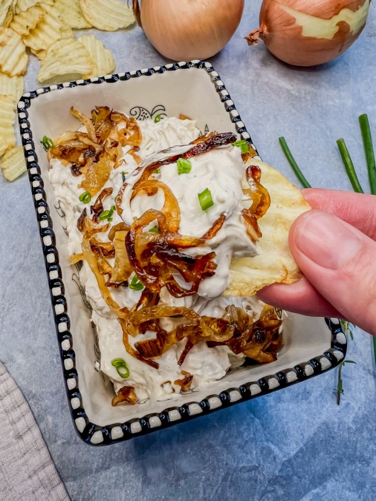 Caramelized french onion dip in a glass dish as some is being scooped out with a chip