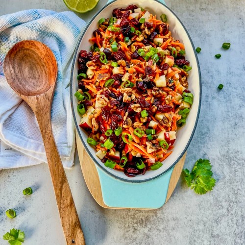 Carrot apple walnut salad in a serving dish with a spoon next to it and some fresh herbs scattered around