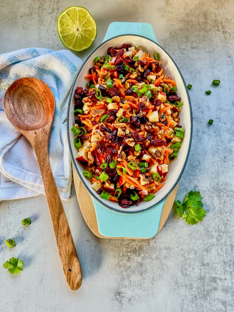 Carrot apple walnut salad in a serving dish with a spoon next to it and some fresh herbs scattered around