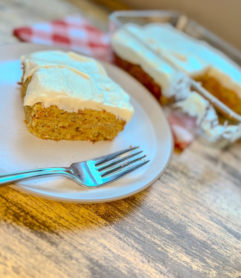 Carrot cake bars in a glass pan with a white plate in front of it with a carrot cake bar on it