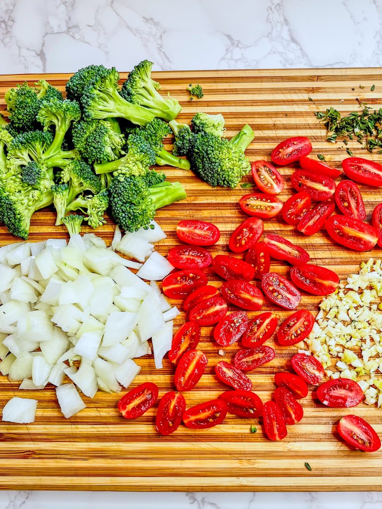 Tomato, onion, garlic, broccoli, and thyme cut up on a wooden cutting board