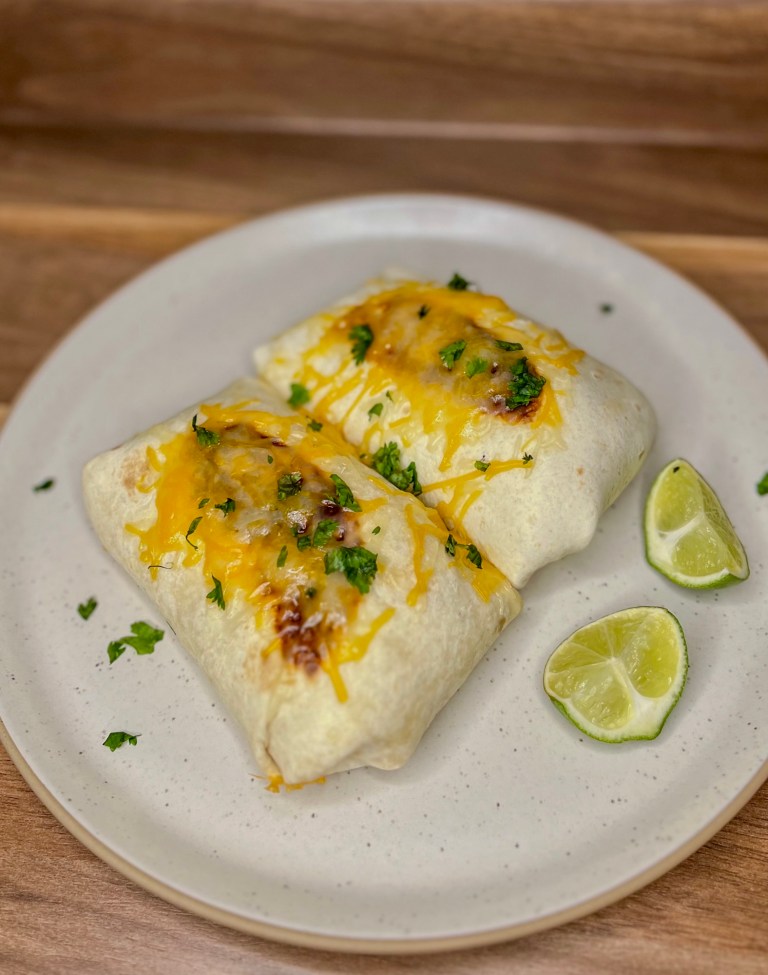 Two cheesy chicken burritos with melted cheese on the top on a white plate with some cilantro and lime wedges