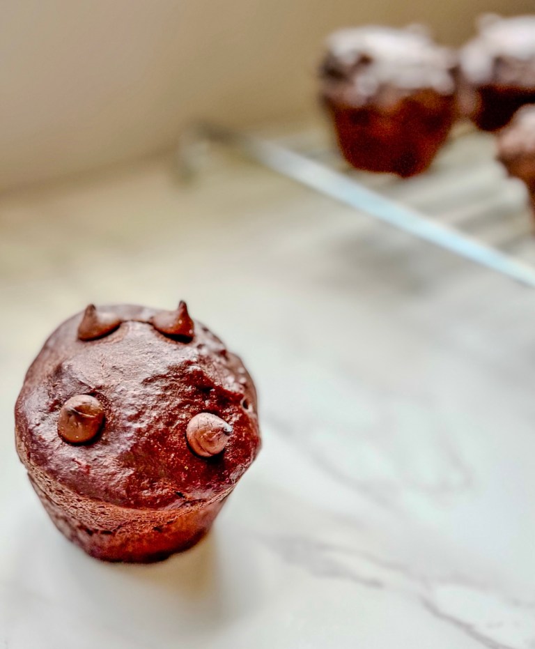 Chocolate banana muffins on a countertop with one sitting in front and the rest on a cooling rack behind
