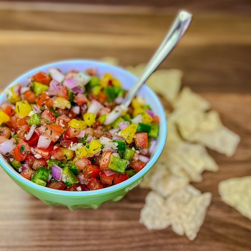 Chunky homemade salsa in a green bowl with a spoon sticking out of it and some chips scattered around