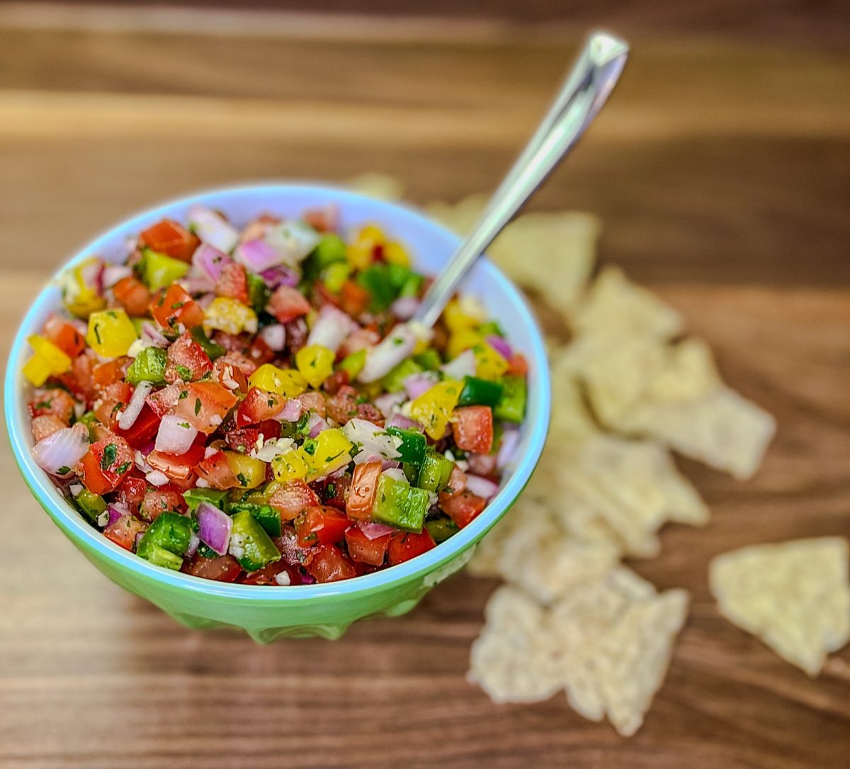 Chunky homemade salsa in a green bowl with a spoon sticking out of it and some chips scattered around