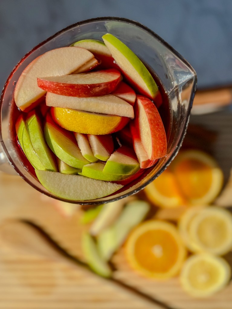 Apple, orange, and lemon in a large pitcher of sangria with fruit scattered nearby
