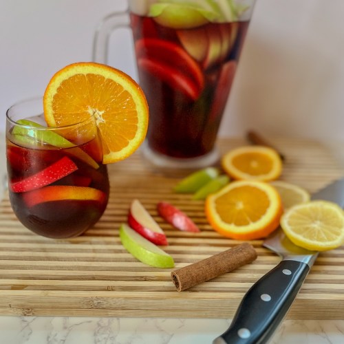 Cranberry sangria in a glass with a bunch of fresh fruit scattered around with a large pitcher filled behind it