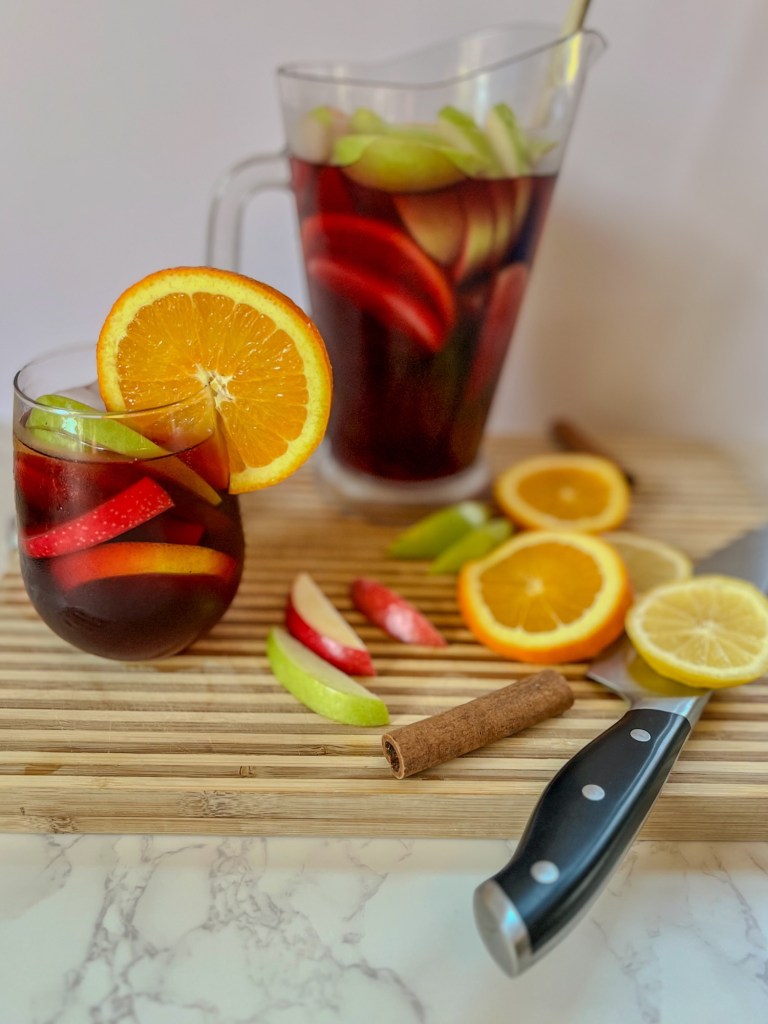 Cranberry sangria in a glass with a bunch of fresh fruit scattered around with a large pitcher filled behind it