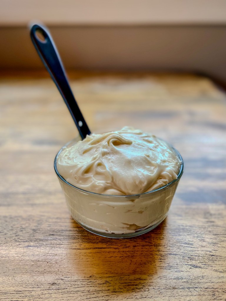 Cream cheese frosting in a glass dish with a rubber spatula sticking out of it, on a wooden table