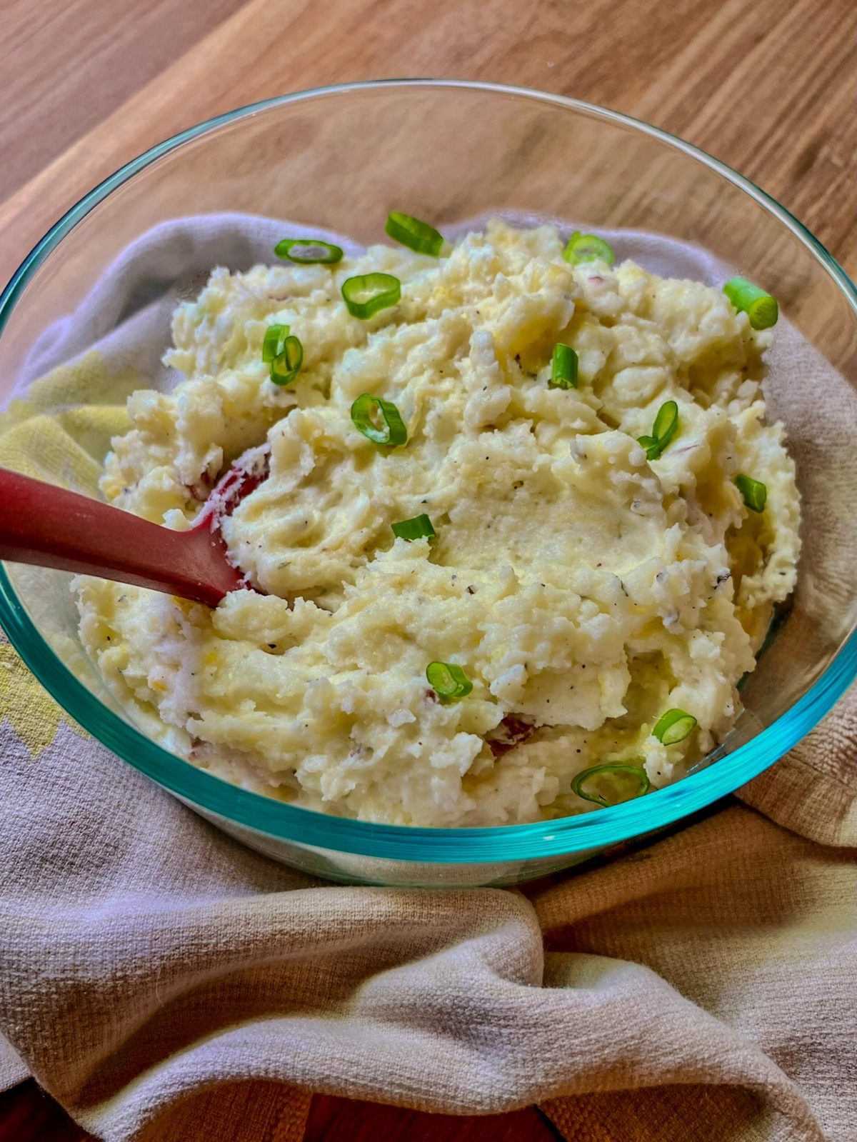 Creamy mashed potatoes in a glass dish with green onion on top and a red serving spoon inside