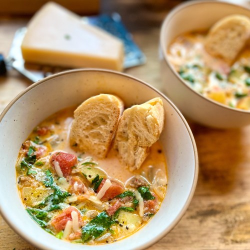 A bowl filled with creamy sausage and potato soup with some baguette slices in it with a large pot of it behind