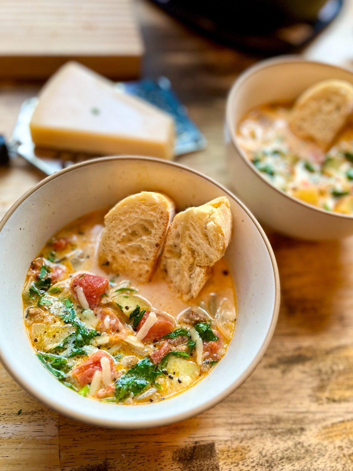 A bowl filled with creamy sausage and potato soup with some baguette slices in it with a large pot of it behind