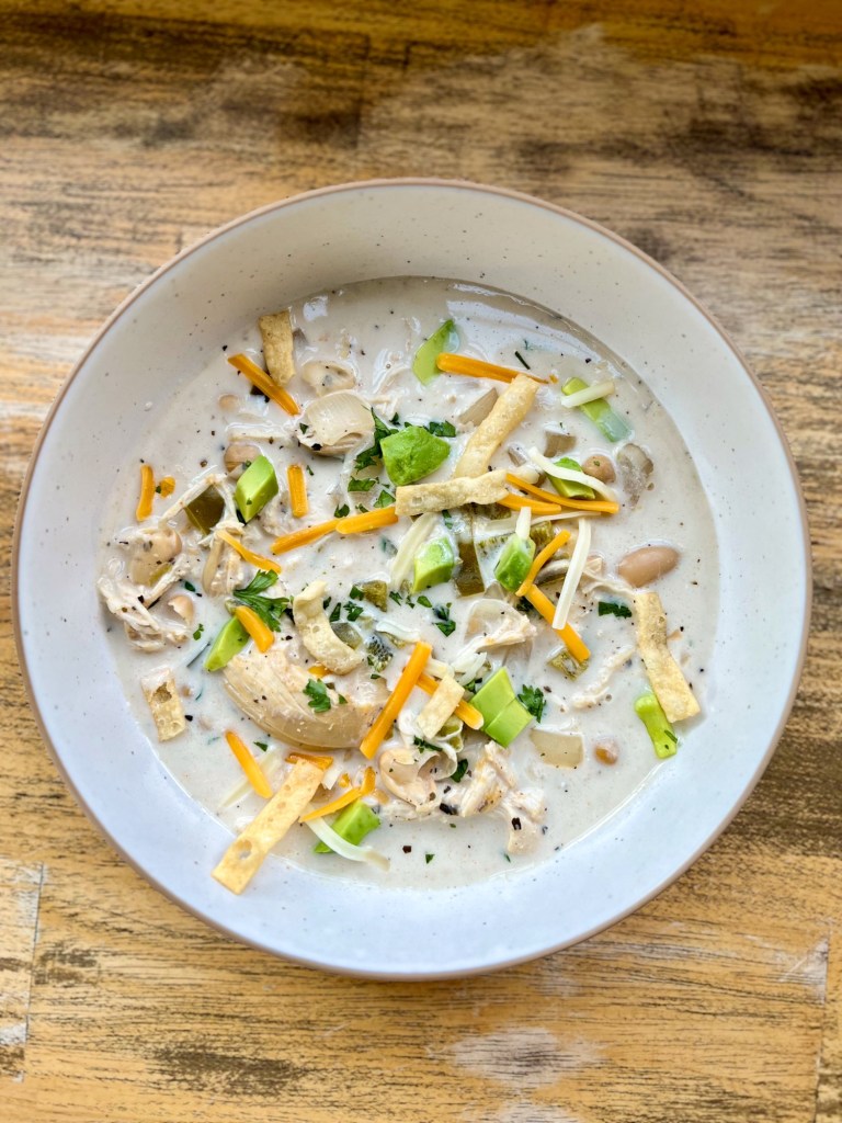 Crockpot white chicken chili in a white bowl with cheese, cilantro, wonton strips, and avocado garnished on top
