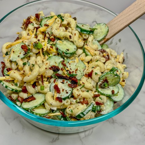 Cucumber Bacon Pasta Salad in a glass dish with a wooden spoon on a marble countertop