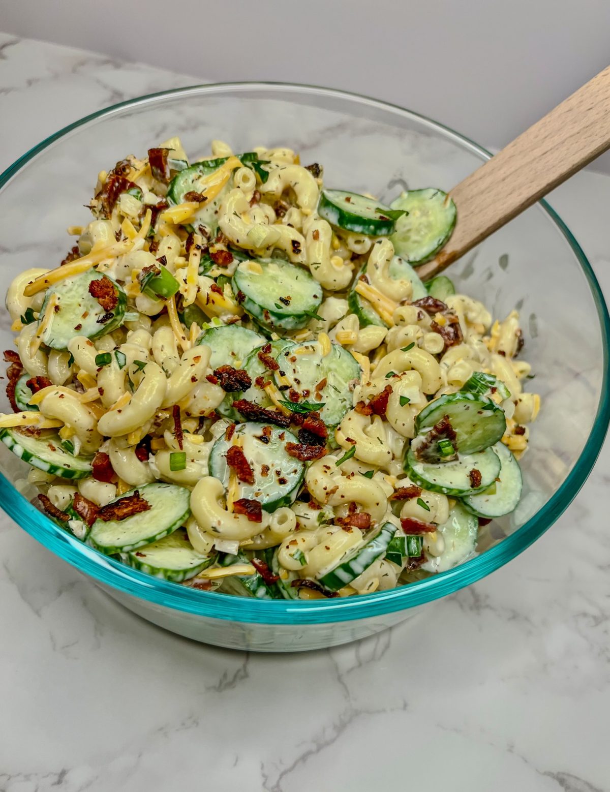 Cucumber Bacon Pasta Salad in a glass dish with a wooden spoon on a marble countertop