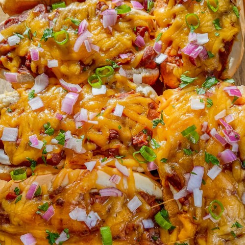 Chili cheese dogs in a glass baking dish with garnishes on top on a wooden table