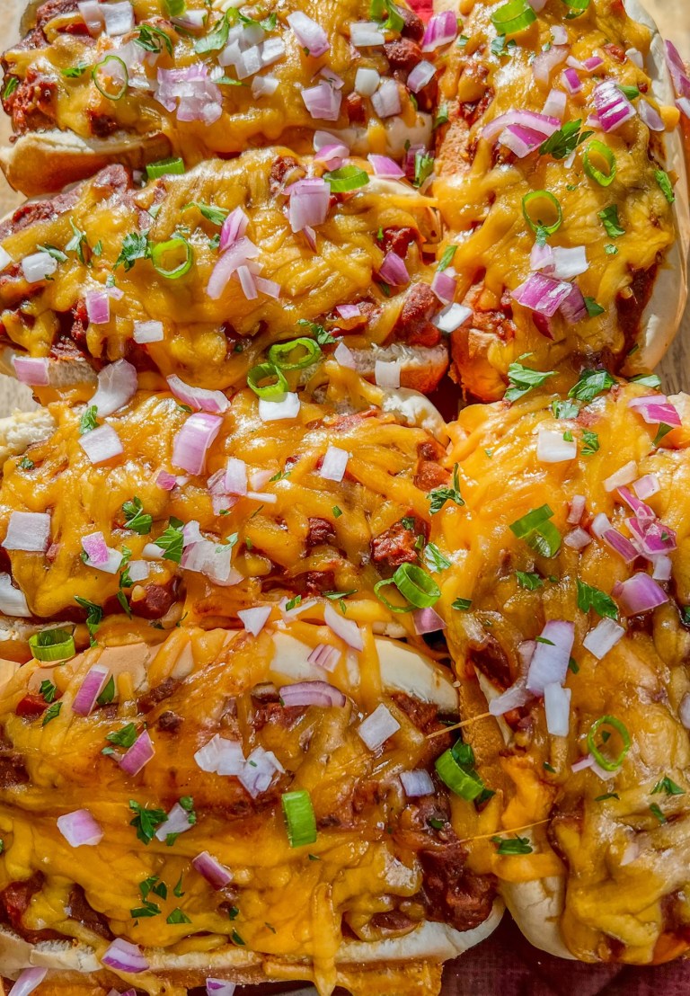 Chili cheese dogs in a glass baking dish with garnishes on top on a wooden table