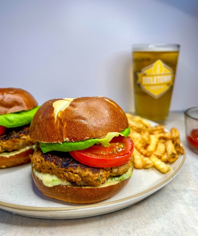 Green onion turkey burgers on a pretzel bun with tomato, lettuce, and honey mustard mayo on a plate with some fries, ketchup, and beer next to it