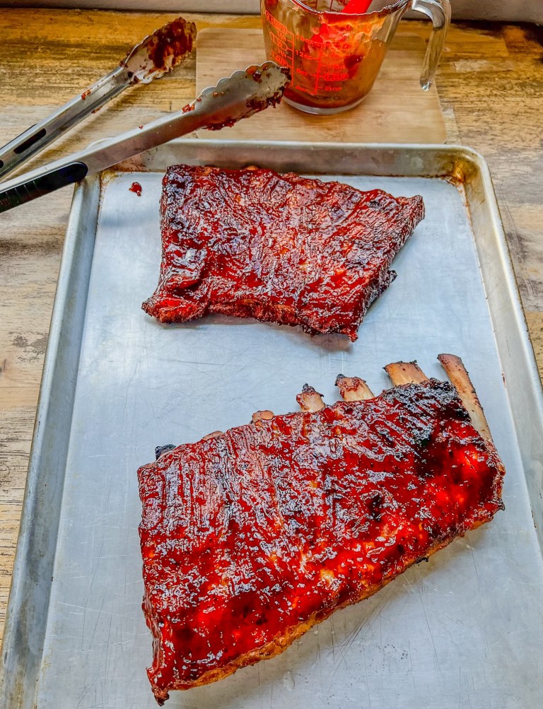 Grilled BBQ Ribs on a baking sheet with some sauce and tongs nearby