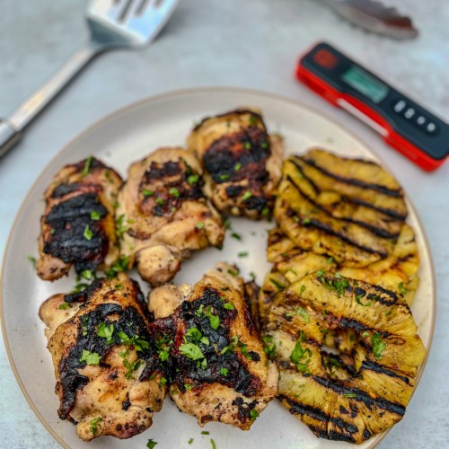 Hawaiian chicken thighs and grilled pineapple on a white plate with a spatula, tongs, and meat thermometer nearby