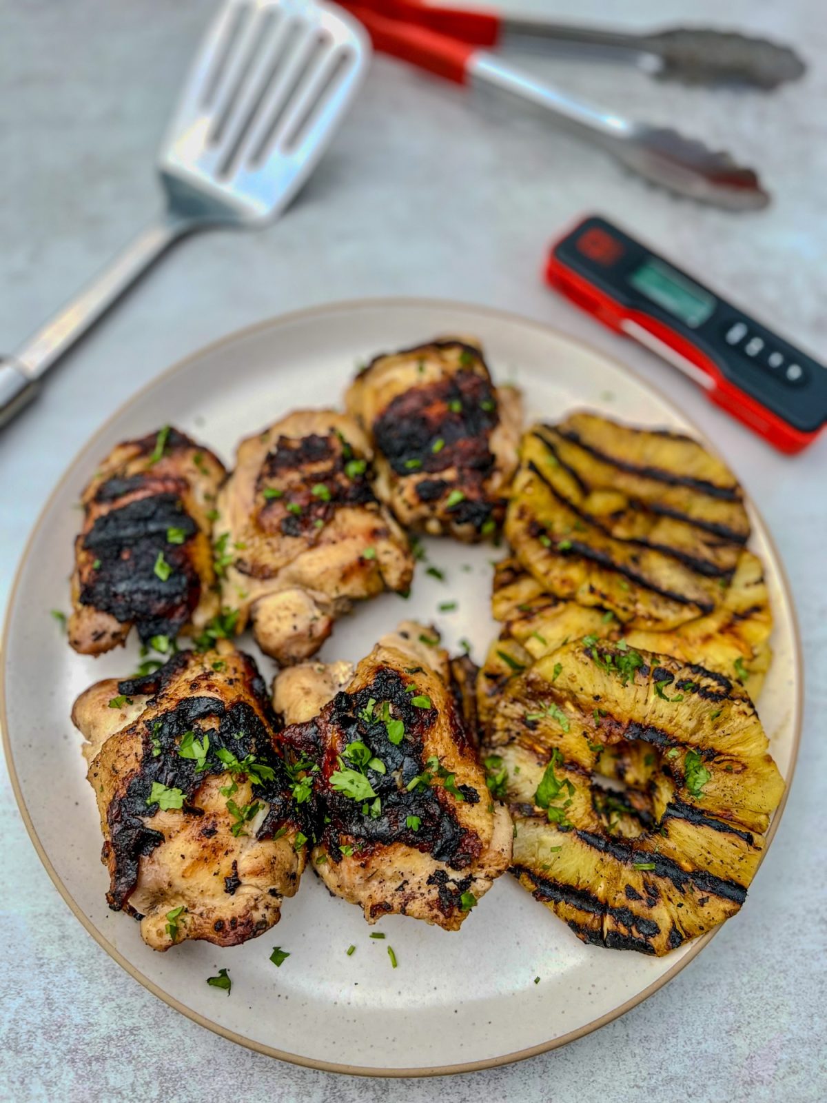 Hawaiian chicken thighs and grilled pineapple on a white plate with a spatula, tongs, and meat thermometer nearby