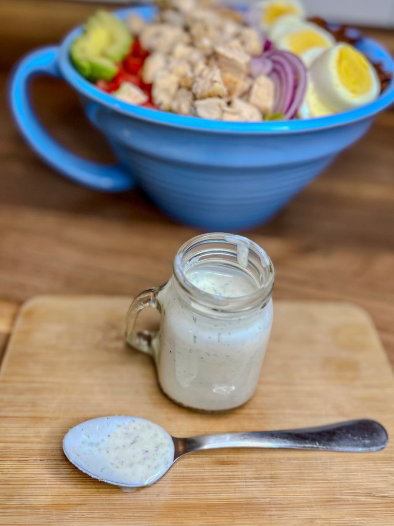 Homemade ranch in a small glass jar with a spoonful of it in front of it and a big salad behind