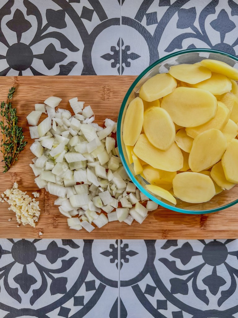 Sliced potatoes, diced onion, minced garlic, and thyme on a wooden cutting board