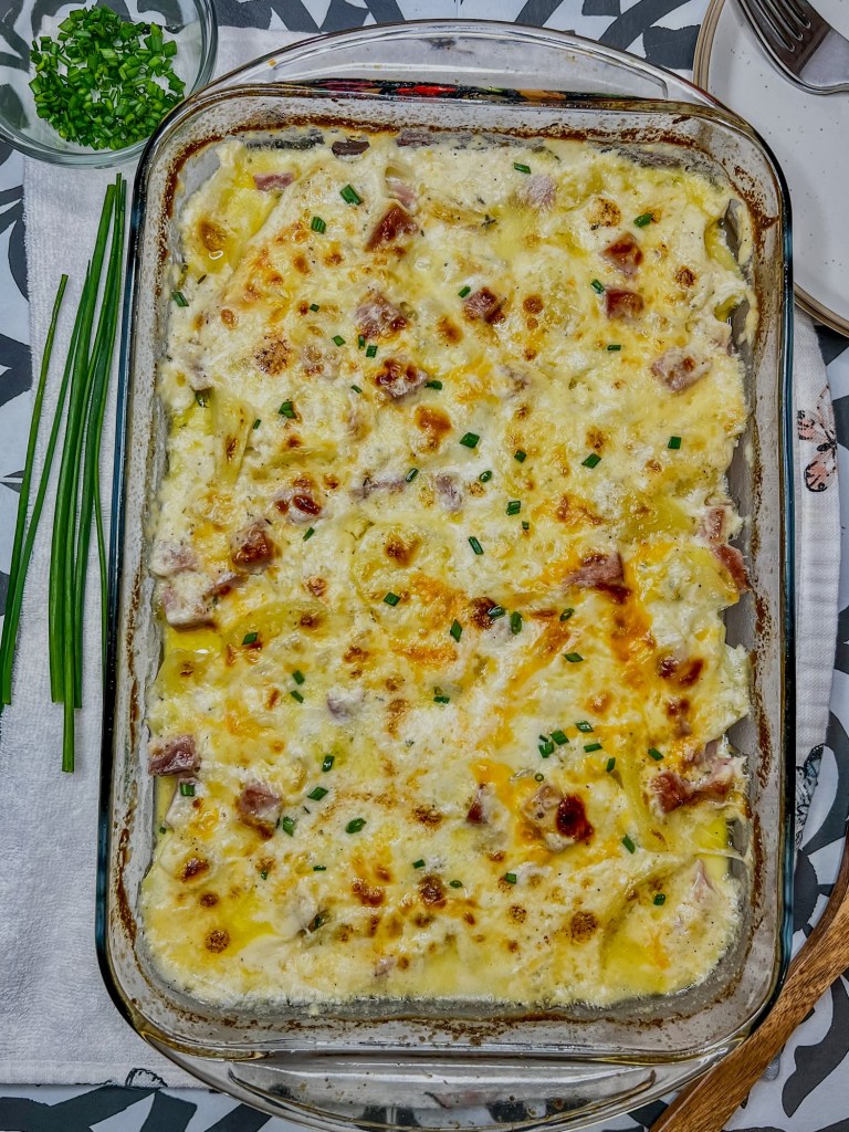 Baked ham and scalloped potatoes in a baking dish with a wooden spoon and some herbs nearby