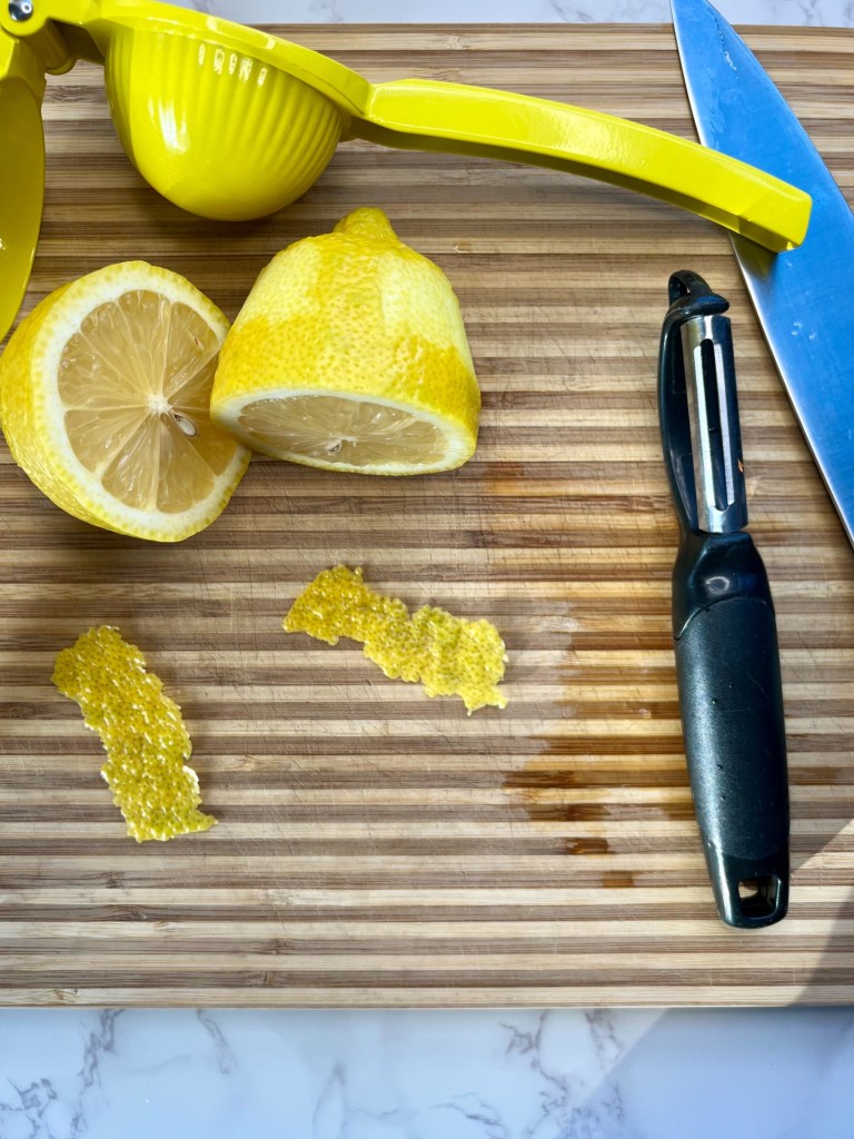 A lemon cut in half on a cutting board with 2 slices of lemon zest peeled off and set to the side