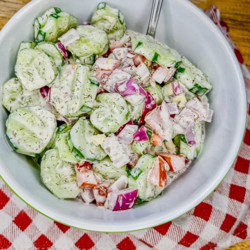 Creamy cucumber salad in a bowl with a red and white towel underneath it on a wooden table