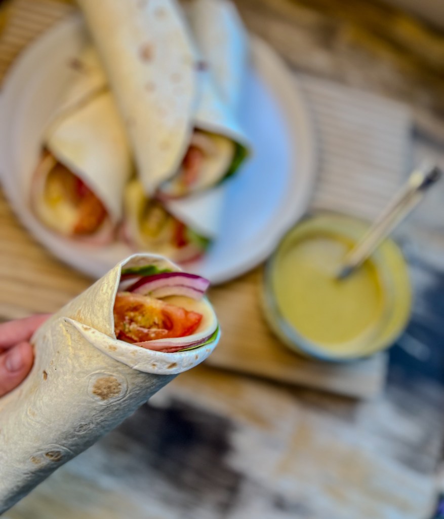 A hand holding an italian wrap over a table with a plate of the wraps and a dish filled with dressing behind it