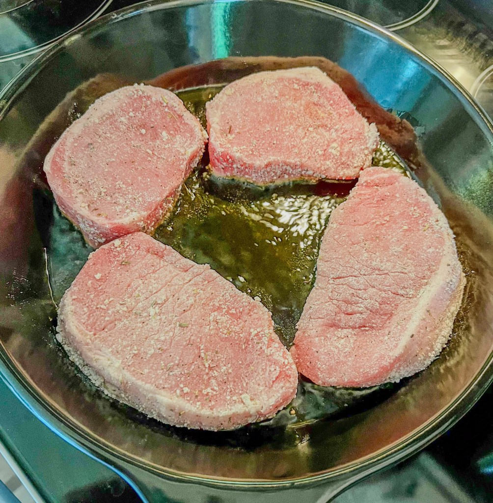 Four pork chops that are lightly coated being seared in a pan on the stove