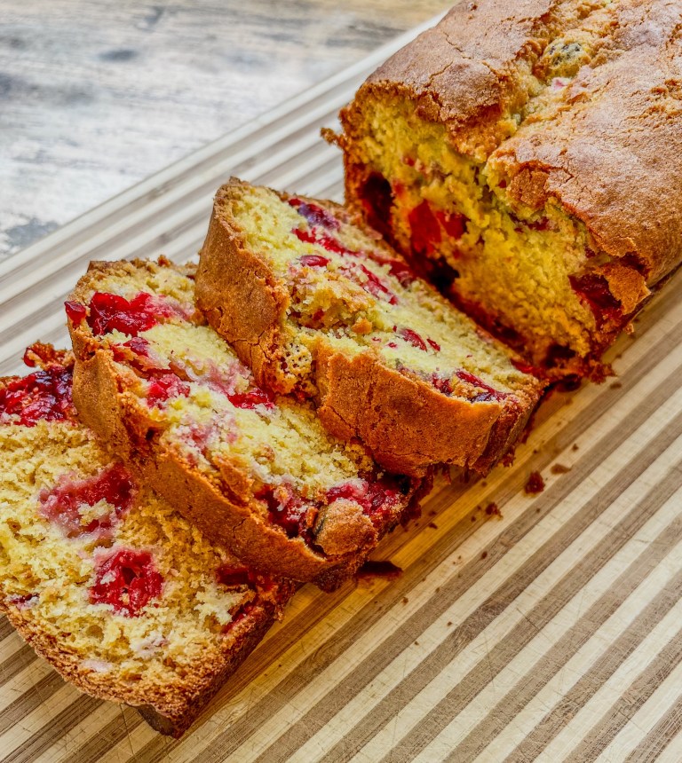 A cranberry orange bread loaf that's been sliced, on a wooden cutting board