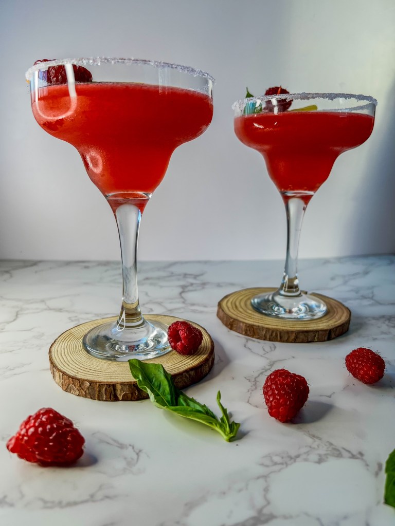 Raspberry honey lemon daiquiri drinks on coasters with herbs and garnishes scattered around it