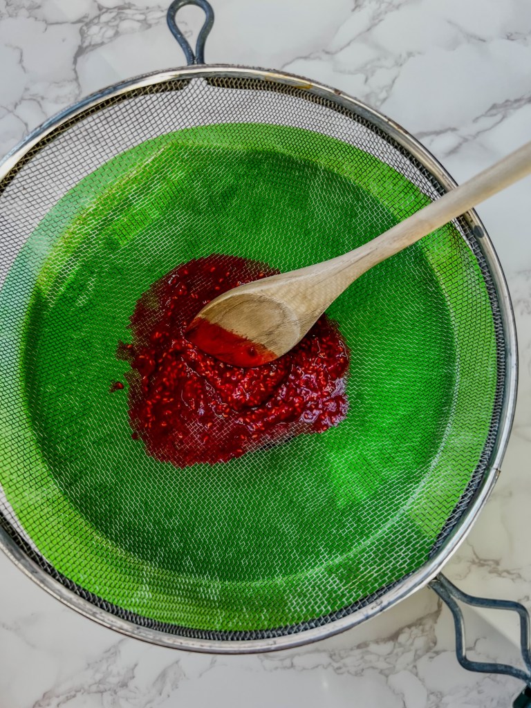 Raspberry puree being strained through a fine mesh sieve into a mixing bowl