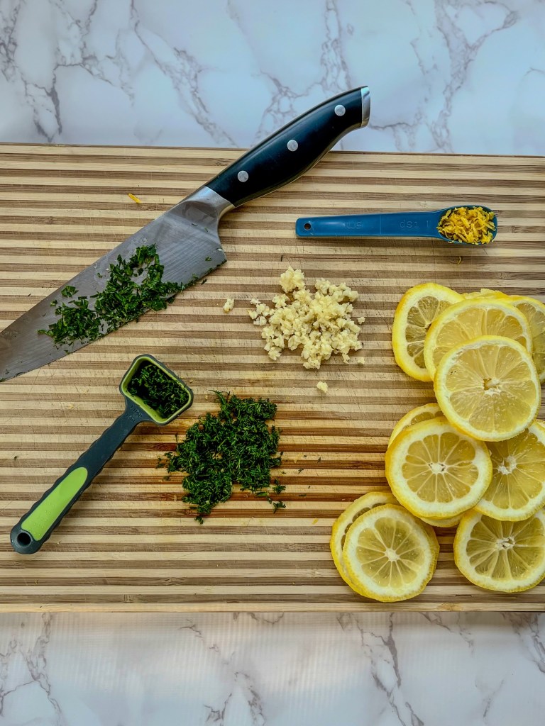 Lemon, dill, garlic, and parsley cut up on a wooden cutting board