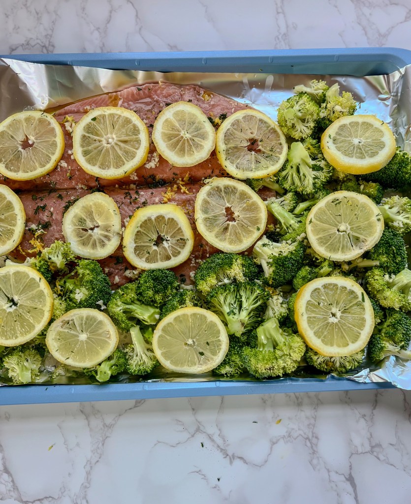 Salmon and broccoli on a baking sheet lined with foil with lemons, garlic, and herbs sprinkled around