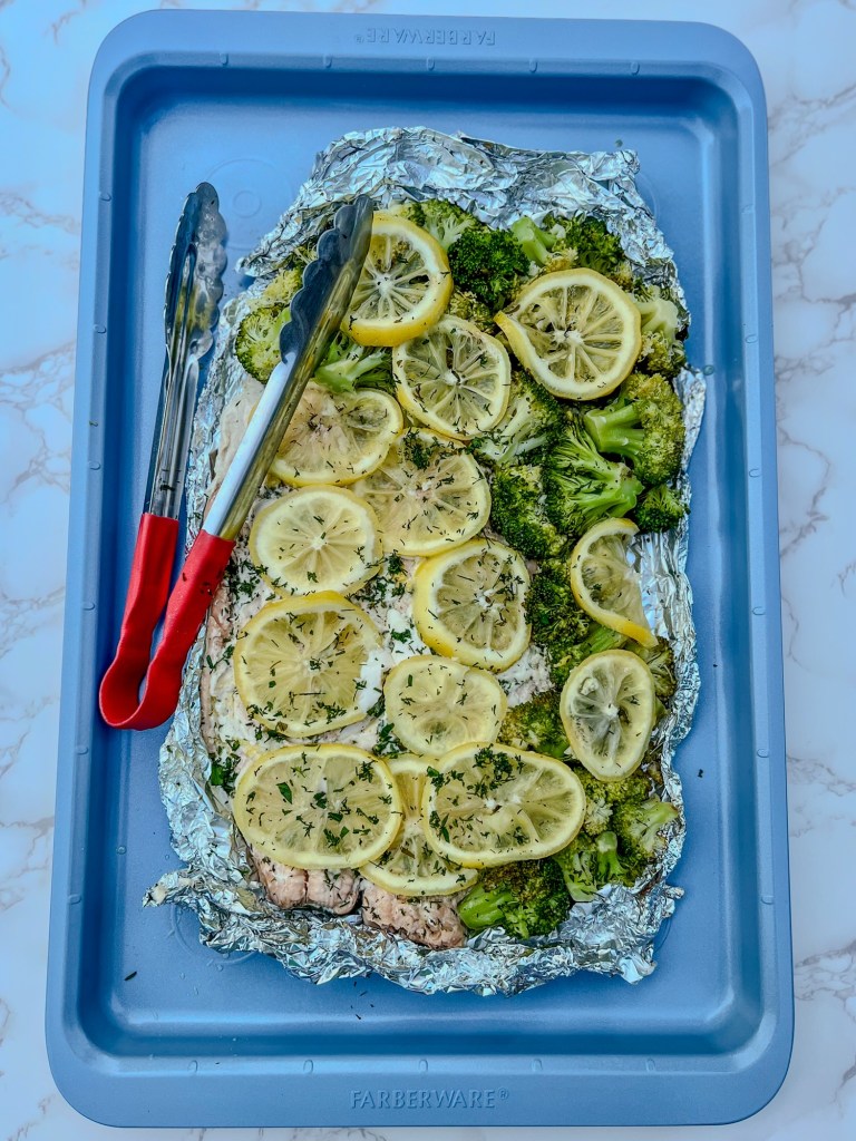 Salmon and broccoli foil pack opened up on a blue baking sheet after just being grilled