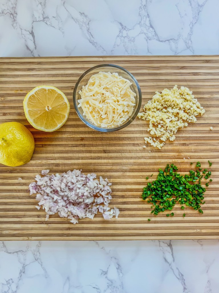 Lemon, shallot, garlic, and chives cut up on a cutting board with some shredded parmesan next to it