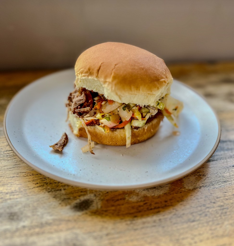 A Smoked Pulled Pork Sandwich with slaw on it on a white plate on a wood table