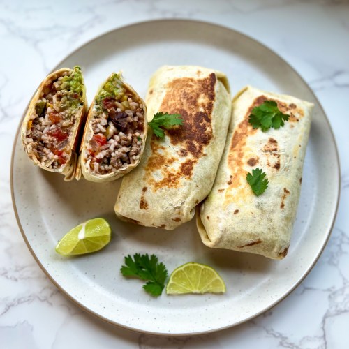 Three southwest burritos with one cut in half on a white plate with lime and cilantro on the side
