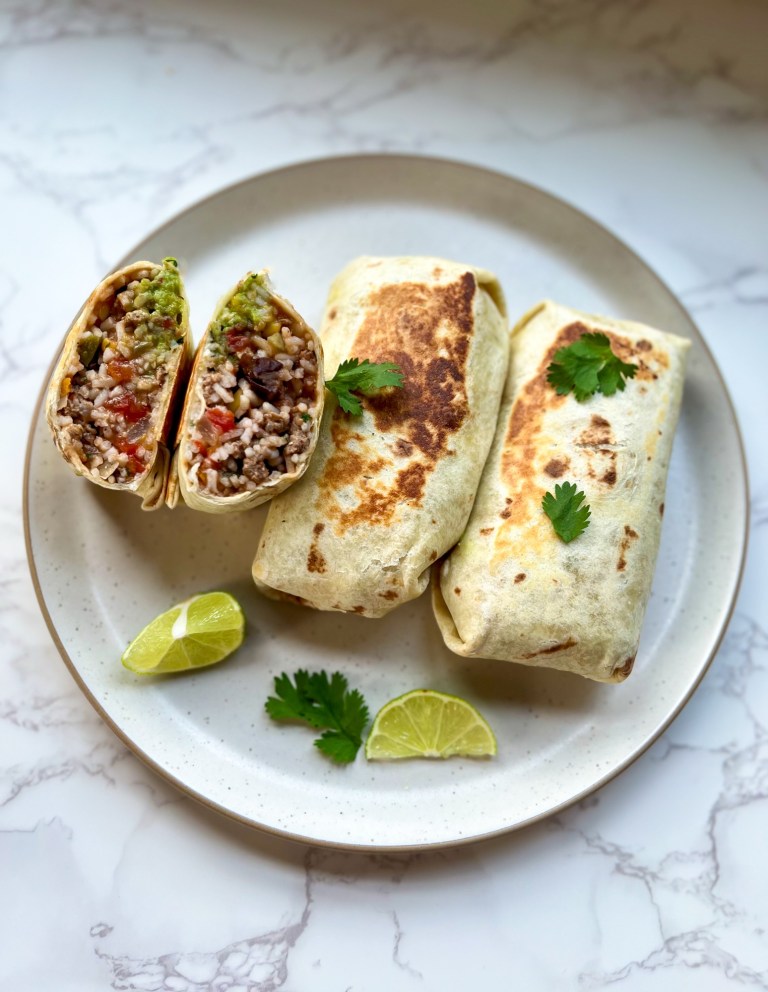 Three southwest burritos with one cut in half on a white plate with lime and cilantro on the side