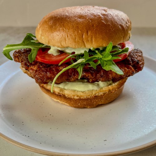 A spicy fried chicken sandwich with tomato, arugula, and avocado blue cheese dressing on a white plate