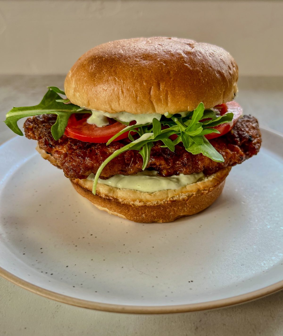 A spicy fried chicken sandwich with tomato, arugula, and avocado blue cheese dressing on a white plate