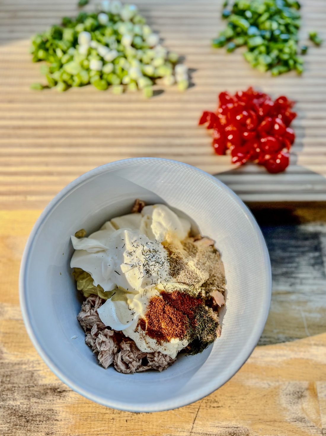 A bowl filled with ingredients to make pasta salad with cut up veggies behind it on a wooden cutting board